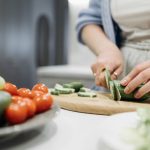 Close-up of a woman slicing cucumbers on a wooden board surrounded by fresh vegetables.