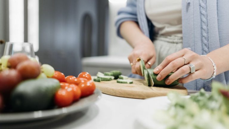 Close-up of a woman slicing cucumbers on a wooden board surrounded by fresh vegetables.